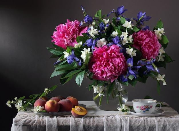 Blumenstrauß aus Pfingstrosen, Iris und Jasmin im Glas und Pfirsichen auf einem Tisch mit Spitzentischdecke.