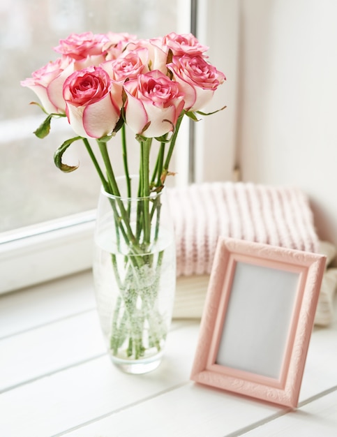 Blumenstrauß aus Rosen und Fotorahmen am Fenster. Valentinskarte. Komposition mit frischen Blumen. Platz für Text. Muttertag und 8. März Karte