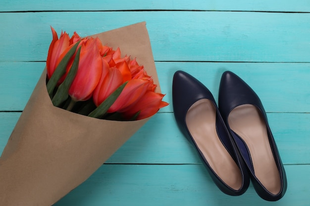 Foto blumenstrauß aus roten tulpen und schuhen mit hohen absätzen auf blauem hintergrund aus holz. feiertag muttertag oder 8. märz, geburtstag. ansicht von oben