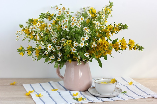 Blumenstrauß aus weißen und gelben Blumen, Lysimachia und Kamille in einem Krug und einer weißen Tasse auf dem Tisch.