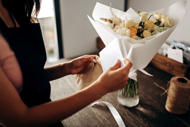 Blumenstrauß in Pastellfarben verpackt in weißem Papier befindet sich in einer Vase. Florist steckt eine Postkarte in einen Umschlag.