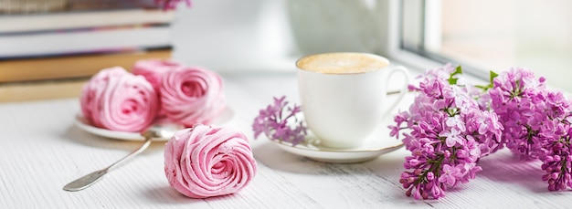 Blumenstrauß von Fliedern, von Tasse Kaffee, von selbst gemachtem Eibisch und von Stapel Büchern auf Fensterbrett Romantischer Frühlingsmorgen.