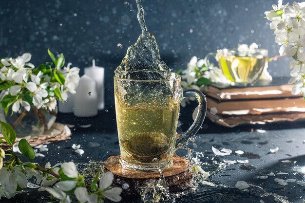 Blumentee in einem transparenten Becher auf dunklem Hintergrund. Neben der Tasse sind Bücher und Zweige mit weißen Apfelblüten. Der Tee schwappt aus der Tasse