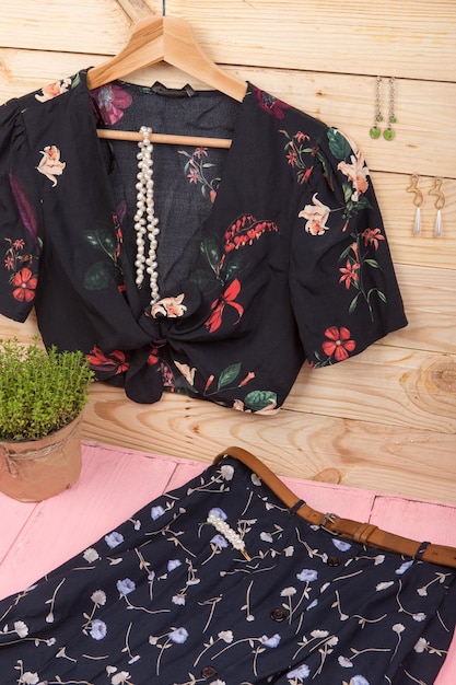 Blusa preta com estampa floral pendurada no cabide cinto de saia azul e brincos de colar de pérolas de cabelo jóias em fundo de madeira