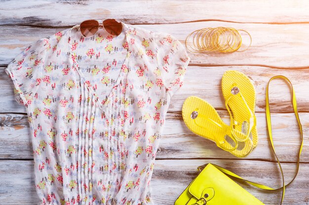 Bluse mit V-Ausschnitt und Flip-Flops. Gelbe Schuhe und Armbänder der Frau. Kleidung auf weißem Holzboden. Verkleiden Sie sich für den Sommer.