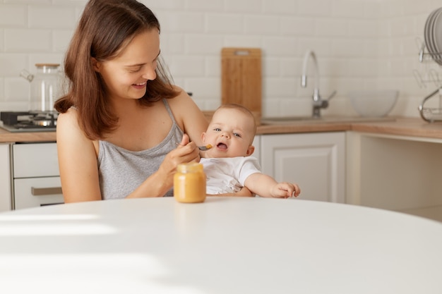 Boa aparência feliz mãe sentada com a filha ou filho nas mãos à mesa e alimentando o bebê com purê do pote, tiro interno da mamãe e comendo a criança.