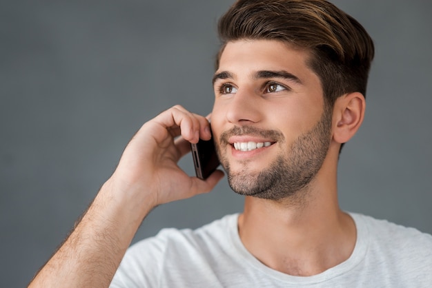 Boa conversa com o amigo. Jovem feliz falando no celular e sorrindo em pé contra um fundo cinza