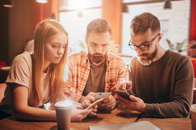 Boa foto de três amigos concentrados sentados juntos à mesa e olhando para seus telefones. Eles estão jogando jogos.