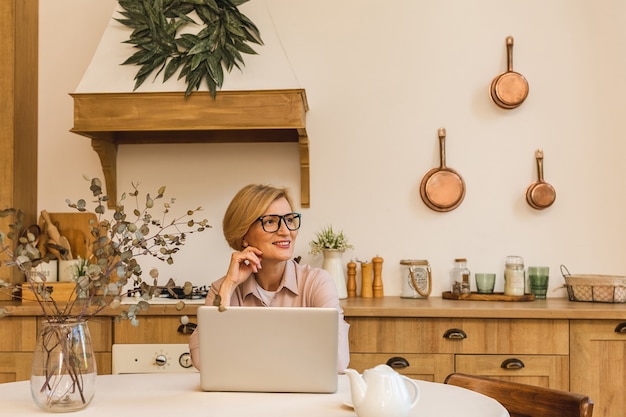 Boa manhã. Alegre sorridente mulher idosa sênior em pé na cozinha e usando seu laptop enquanto descansava depois do café da manhã, freelancer, trabalhando em casa.