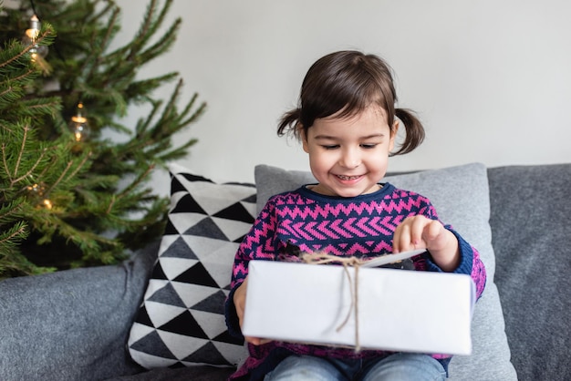 Boas festas e feliz natal menina bonitinha com presente de natal garoto aproveite o feriado e abra o presente sentado no sofá cinza na sala