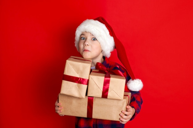 Boas festas e Feliz Natal. Retrato de um menino em um boné com presentes nas mãos sobre um fundo vermelho. Um lugar para texto. Foto de alta qualidade