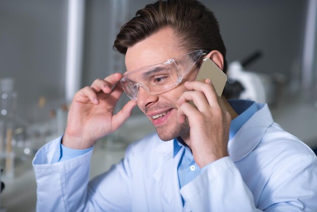 Boas notícias Agradável e bonito praticante inabalável em óculos especiais e uniforme sentado na sala de mãos dadas perto da cabeça e falando pelo celular