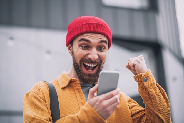 Boas notícias. Jovem barbudo com um chapéu vermelho olhando para o telefone e se sentindo feliz