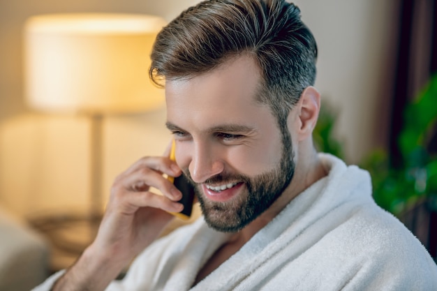 Boas notícias. Jovem barbudo com um roupão de banho branco falando ao telefone e parecendo satisfeito