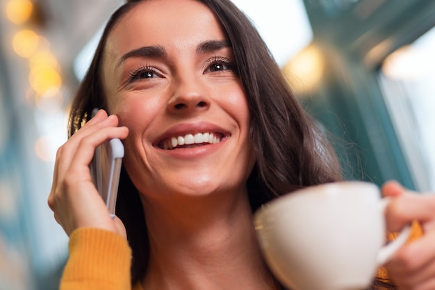 Boas notícias. perto de uma atraente mulher morena feliz segurando o telefone enquanto sorri e bebe chá