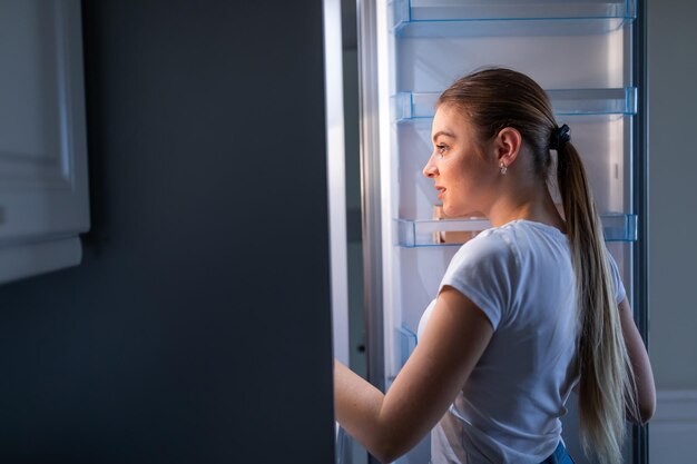 Bocado nocturno Alegre mujer madura abriendo el refrigerador y posando de perfil