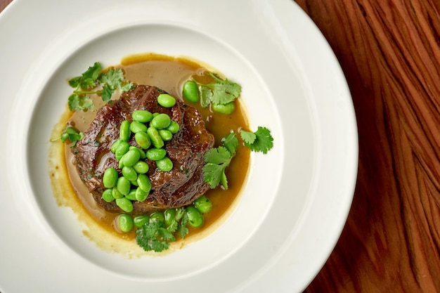 Bochecha de carne estufada com molho de cogumelos e feijão edamame num prato branco sobre fundo de madeira