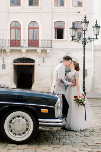 Boda de lujo feliz pareja besándose y abrazándose cerca del coche retro negro en el centro antiguo de la ciudad, edificios antiguos en el fondo.