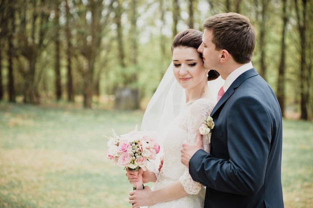 Boda en la primavera. El novio besa y abraza a la novia. Vestido blanco. Un ramo de rosas.