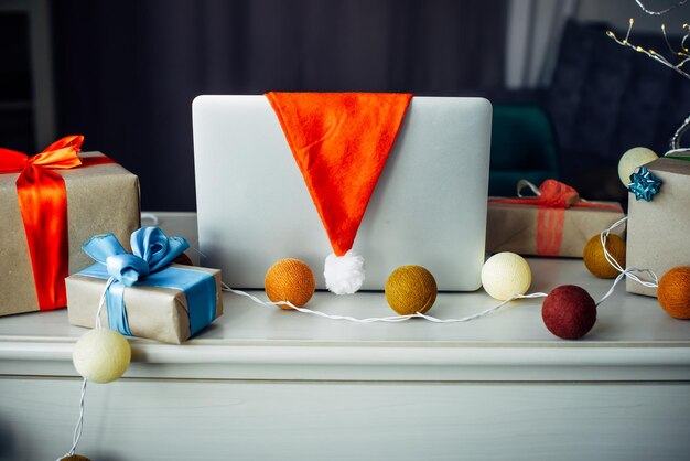 Bodegón de año nuevo en el interior de una casa, primer plano. El gorro de Papá Noel cuelga de un portátil, cajas de regalo y una guirnalda de bolas de colores. Tarjeta de Navidad con espacio de copia.