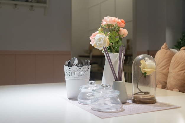 Bodegón de comedor. Ciérrese para arriba de la mesa de comedor adornada con las flores color de rosa en pequeño vidrio y florero con la cocina en fondo.