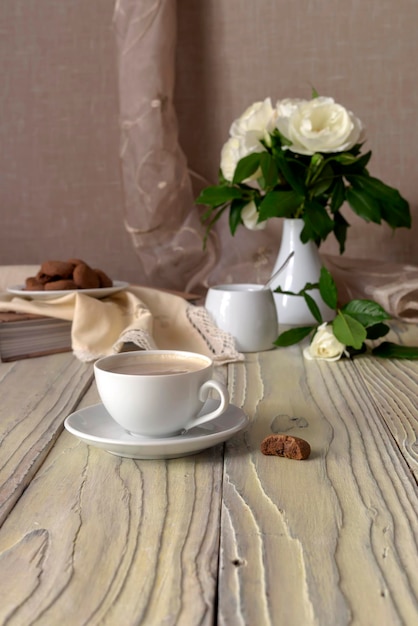 Bodegón Copa con ramo de capuchino de rosas blancas y galletas de chocolate en un primer plano de la mesa de madera