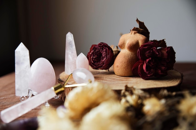 Bodegón con una figura de barro de una mujer embarazada decorada con rosas secas y varios cristales de cuarzo rosa Rodillo de cara de huevo de cuarzo rosa