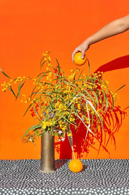 Bodegón de flores de primavera con sombra dura y una mano con una naranja.