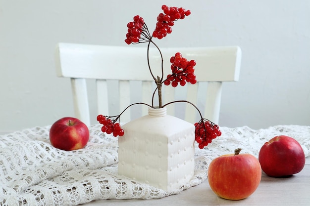 Bodegón de invierno jarrón blanco con ramas rojas de manzanas rojas viburnum en una servilleta blanca en una servilleta blanca contra una silla