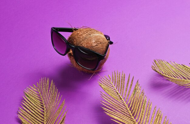 Bodegón de moda creativa. Coco con gafas de sol con hojas de palma doradas sobre fondo morado