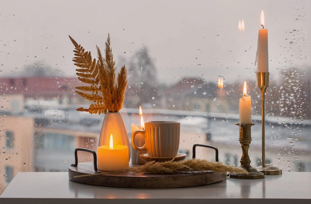 Bodegón de otoño con velas encendidas y una taza de café en el fondo de una ventana con raindr