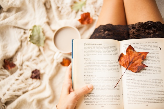 Bodegón de otoño. Vista desde arriba. La niña lee un libro abierto mientras está sentada en una tela escocesa