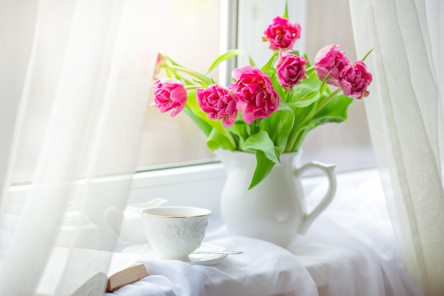 Un bodegón primaveral, un ramo de tulipanes en un jarrón, una taza de té y un libro antiguo, yacen en la ventana