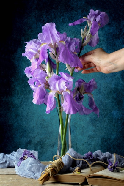 Bodegón con ramo de lirios morados dentro de un jarrón de vidrio sobre una mesa rústica con muselina en libro vintage azul y cuerda con la mano recogiendo una flor