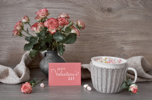 Bodegón de San Valentín con taza de chocolate caliente con malvaviscos, rosas rosadas y tarjeta de felicitación con texto
