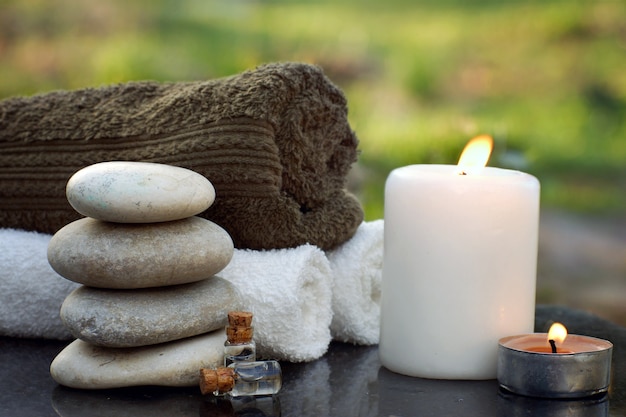 Bodegón de spa con toallas, una vela encendida, aceite de baño y piedras de masaje con el telón de fondo de un jardín verde en verano