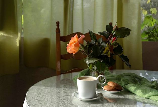 Bodegón con una taza de galletas de capuchino matutino y rosa naranja fresca en un vaso en el primer plano de la mesa