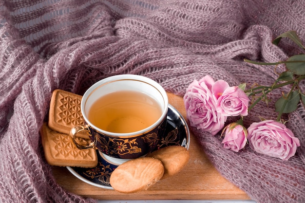 Bodegón de una taza de té, platillo, galletas con rosas rosadas sobre una mesa de madera con un paño de lana.