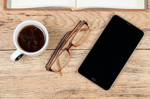 Bodegón de un teléfono inteligente, vasos, taza de café y libro en la mesa de madera vieja