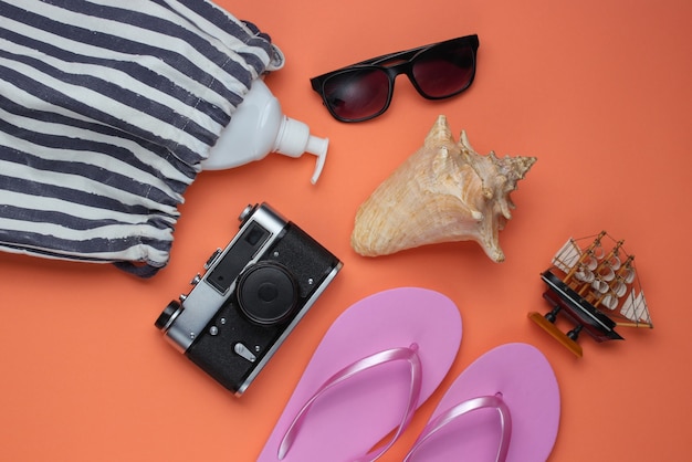 Bodegón de verano. Accesorios de playa. Chanclas rosa de moda, bolso, cámara retro, botella de bloqueador solar, gafas de sol, concha sobre papel coralino.