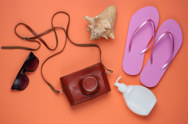Bodegón de verano. Accesorios de playa. Chanclas rosa de moda, cámara retro, botella de bloqueador solar, gafas de sol, concha sobre fondo de papel coral.