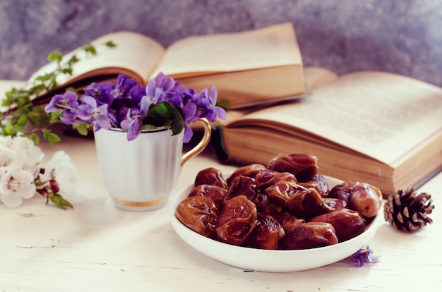 Bodegón con violeta en taza blanca, libros antiguos y frutas de fecha en un plato. Floral romántico