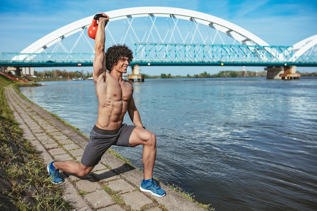 Bodybuilder mit Sixpack, perfektem ABS, Schultern, Bizeps, Trizeps und Brust macht ein starkes Körpertraining mit Kettlebell, in der Nähe des Flusses.