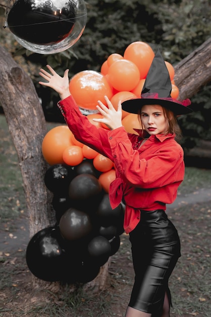 Böses schönes Mädchen in orangefarbenem Hemd, schwarzem Rock und Hexenhut wird ein Ballon-Halloween-Party-Kunstdesign aufwerfen