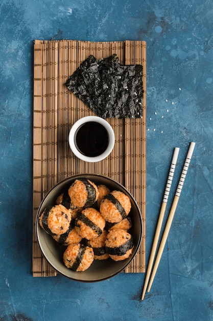 Un bol de bolas de arroz con semillas de sésamo y alga nori, servido con salsa de soja, palillos sobre un fondo azul oscuro. Vista lateral con espacio de copia. Orientación vertical