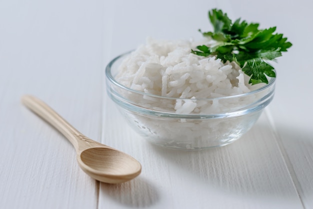 Bol de vidrio con arroz de grano largo y perejil y cuchara de madera en la mesa de madera blanca.