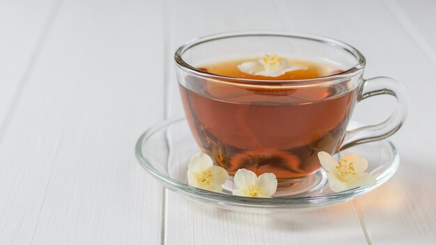 Bol de vidrio con té de hierbas con flores de jazmín en una mesa de madera blanca.