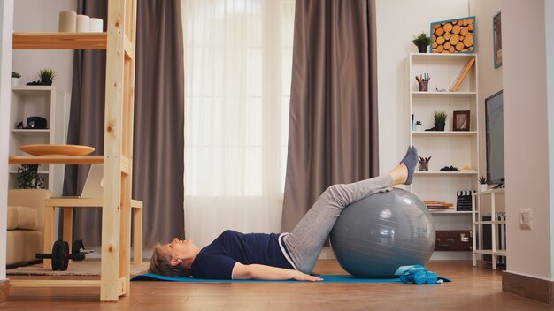 Bola de fitness de treinamento de mulher sênior deitada no tapete de ioga na sala de estar. idoso estilo de vida saudável pensionista vivendo esporte e treinamento de bem-estar dentro de casa em casa