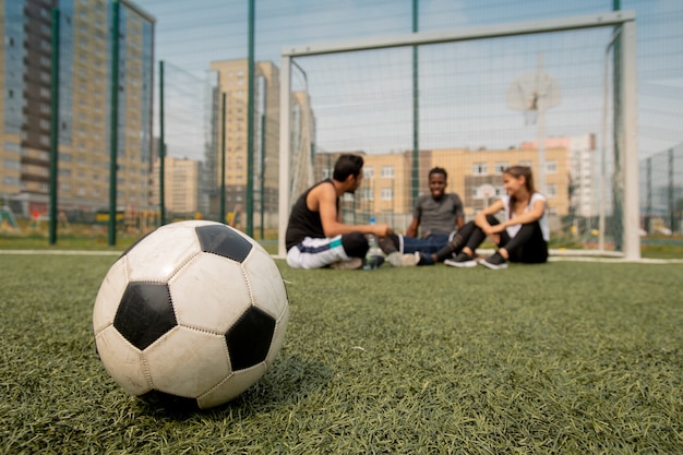 Bola de futebol no campo ou playground de três jovens jogadores fazendo uma pausa após o treino