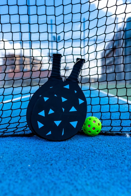 Foto bola de pickleball e pás apoiadas na rede de uma quadra de pickleball